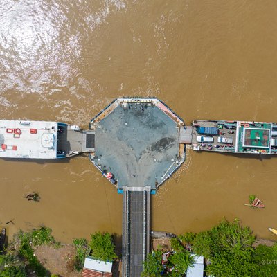 Ảnh chụp từ trên cao cho thấy bến phà nối hai bờ sông với nước sông đục và cây xanh hai bên bờ. Cảnh vật mang lại cảm giác yên bình và tĩnh lặng của vùng đồng bằng sông Cửu Long.