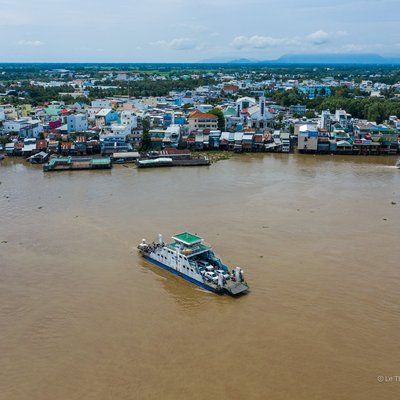Ảnh chụp cảnh một con sông lớn với phà đang di chuyển trên mặt nước, phía sau là khu dân cư đông đúc và cây xanh. Khung cảnh tạo cảm giác yên bình và rộng lớn dưới ánh sáng ban ngày dịu nhẹ.