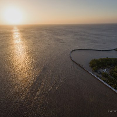 Bức ảnh chụp cảnh hoàng hôn trên vùng nước rộng lớn với một mảnh đất có rừng ngập mặn ở góc phải. Cảnh vật tạo cảm giác yên bình và tĩnh lặng, ánh sáng mềm mại của hoàng hôn làm nổi bật vẻ đẹp tự nhiên của vùng đất này.