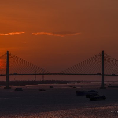 Bức ảnh chụp cảnh hoàng hôn trên cầu Rạch Miễu bắc qua sông lớn với mặt trời lặn rực rỡ phía sau. Toàn cảnh tạo cảm giác yên bình và tĩnh lặng của một buổi chiều cuối ngày trên vùng đồng bằng sông Cửu Long.