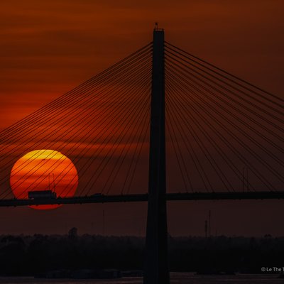 Hình ảnh cầu Rạch Miễu vào lúc hoàng hôn với mặt trời đỏ rực phía sau cầu dây văng. Cảnh vật tạo cảm giác yên bình và tĩnh lặng khi ngày dần kết thúc.