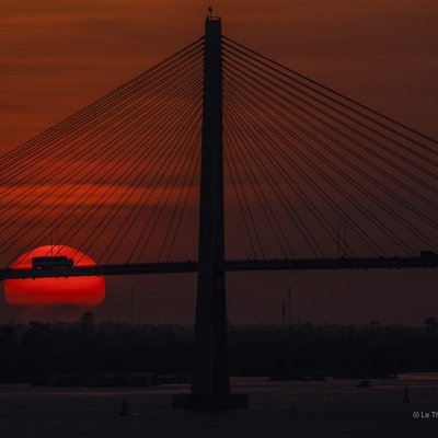 Bức ảnh chụp cầu Rạch Miễu vào lúc hoàng hôn với mặt trời đỏ rực phía sau cầu. Cảnh vật tạo cảm giác yên bình và tĩnh lặng trong không gian đô thị ven sông.