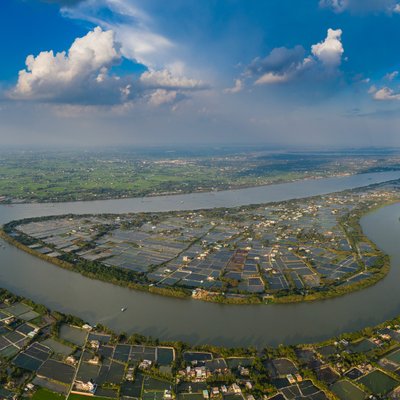 Ảnh chụp toàn cảnh sông Vàm Cỏ với cảnh đồng bằng rộng lớn và các cánh đồng nông nghiệp bao quanh. Cảnh vật mang lại cảm giác yên bình và thanh thản dưới bầu trời xanh với mây trắng.