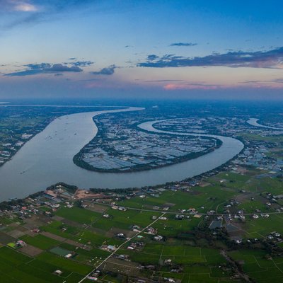 Bức ảnh chụp cảnh sông Vàm Cỏ uốn lượn qua vùng đồng bằng rộng lớn với nhiều cánh đồng và nhà cửa rải rác. Khung cảnh mang lại cảm giác yên bình và rộng mở dưới ánh sáng nhẹ nhàng của buổi chiều.