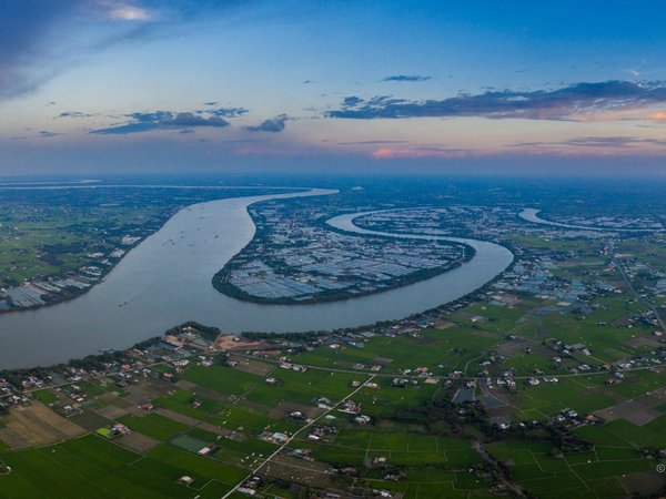 Bức ảnh chụp cảnh sông Vàm Cỏ uốn lượn qua vùng đồng bằng rộng lớn với nhiều cánh đồng và nhà cửa rải rác. Khung cảnh mang lại cảm giác yên bình và rộng mở dưới ánh sáng nhẹ nhàng của buổi chiều.