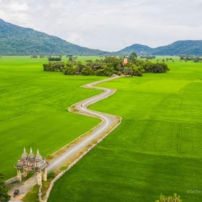 Bức ảnh chụp cảnh đồng lúa xanh mướt với con đường uốn lượn dẫn đến cổng chùa Koh Kas nằm giữa cánh đồng rộng lớn. Khung cảnh mang lại cảm giác yên bình và tĩnh lặng giữa thiên nhiên rộng lớn.