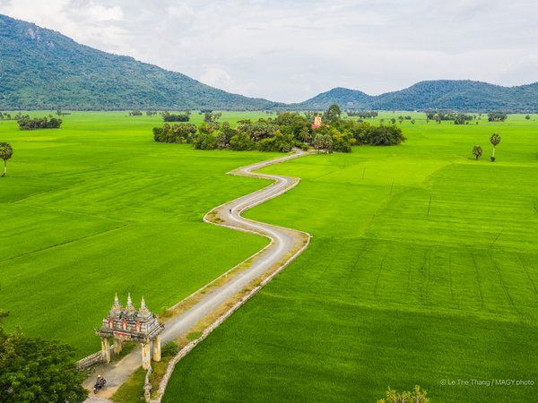 Bức ảnh chụp cảnh đồng lúa xanh mướt với con đường uốn lượn dẫn đến cổng chùa Koh Kas nằm giữa cánh đồng rộng lớn. Khung cảnh mang lại cảm giác yên bình và tĩnh lặng giữa thiên nhiên rộng lớn.