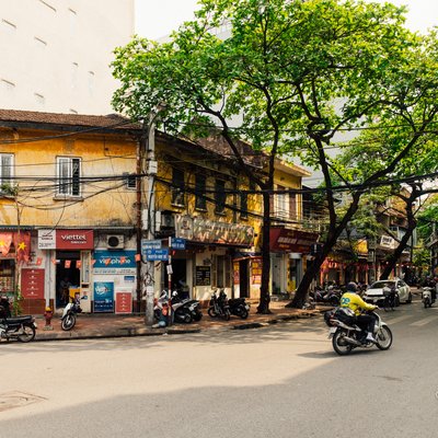 Hình ảnh một góc phố ở Hà Nội với các toà nhà cũ, cây bàng lớn và một vài người đi xe máy trên đường. Không khí yên bình và ánh sáng mặt trời nhẹ nhàng tạo cảm giác thư thái cho cảnh vật.