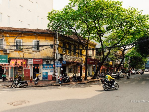 Hình ảnh một góc phố ở Hà Nội với các toà nhà cũ, cây bàng lớn và một vài người đi xe máy trên đường. Không khí yên bình và ánh sáng mặt trời nhẹ nhàng tạo cảm giác thư thái cho cảnh vật.
