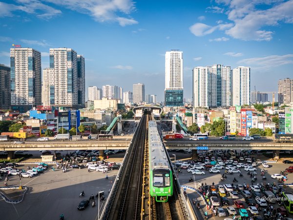 Hình ảnh chụp một nút giao thông đô thị đông đúc với tàu điện trên cao và nhiều phương tiện giao thông dưới đường. Cảnh quan thể hiện sự nhộn nhịp và hiện đại của thành phố vào buổi chiều.