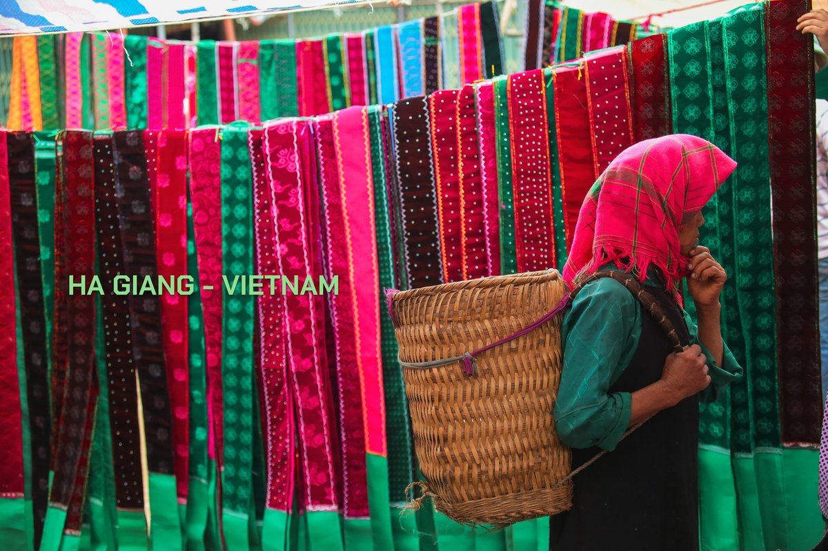 Bức ảnh chụp một người phụ nữ trong trang phục dân tộc tại chợ Hà Giang với các dải vải nhiều màu sắc treo phía sau. Hình ảnh thể hiện sự chân thực và sống động của đời sống thường nhật nơi đây.