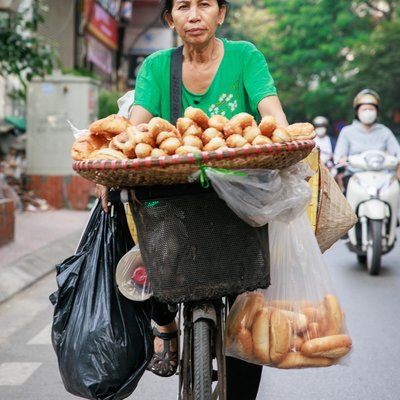 Bức ảnh chụp một người phụ nữ bán hàng rong bánh mì trên chiếc xe đạp ở khu vực đô thị vào buổi sáng. Hình ảnh thể hiện cuộc sống thường nhật bình dị và yên bình của người dân thành phố.