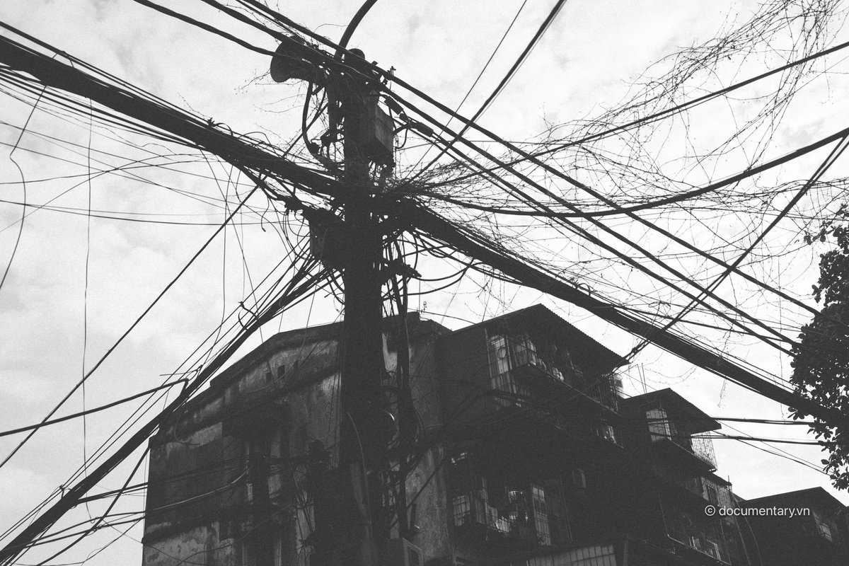Bức ảnh đen trắng chụp cột điện cùng các dây điện chằng chịt trước một toà nhà tập thể cũ ở thành phố. Hình ảnh tạo cảm giác hoài niệm về cuộc sống đô thị giản dị và đời thường.