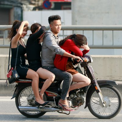 Bức ảnh chụp một nhóm bốn người đang đi trên một chiếc xe máy Honda Dream ở đường phố thành phố. Hình ảnh thể hiện sự đông đúc và nhộn nhịp của giao thông đô thị với cảm giác năng động và vui tươi.