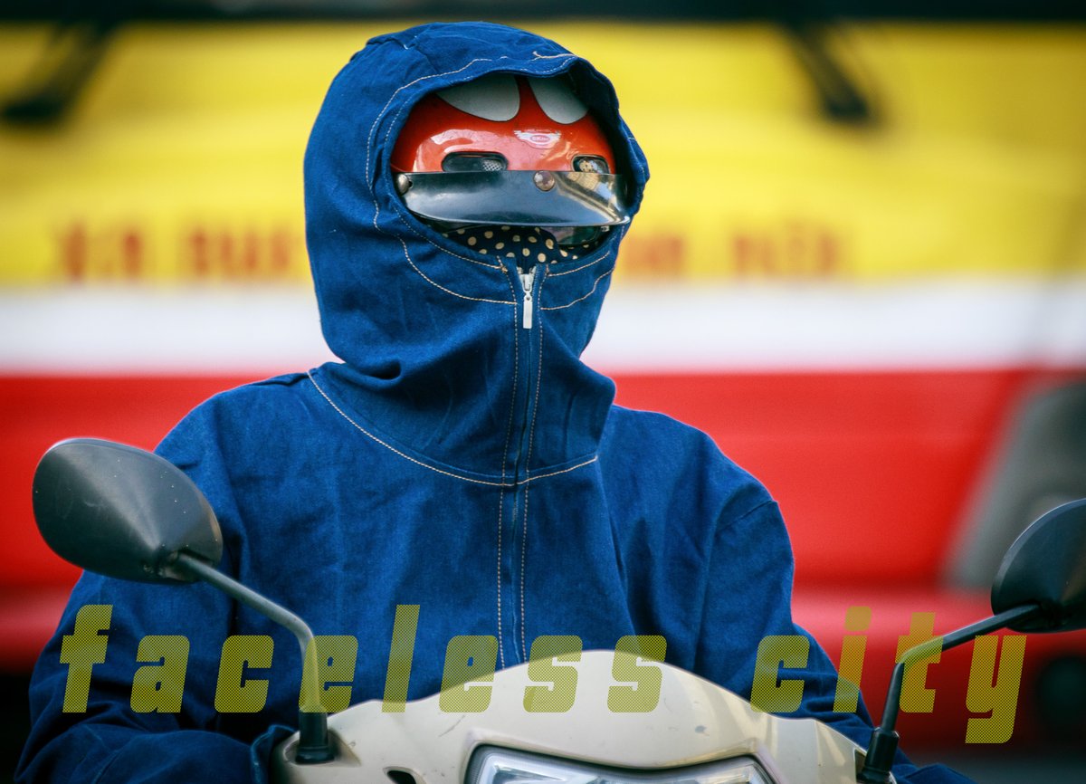 Hình ảnh một người mặc áo chống nắng và mũ bảo hiểm đi xe máy trên đường phố thành phố. Bức ảnh tạo cảm giác cô lập và nhấn mạnh sự vắng mặt rõ ràng của khuôn mặt người trong cảnh đô thị.