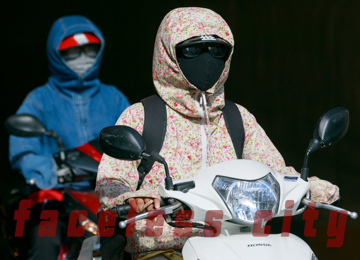 Hình ảnh hai người mặc áo chống nắng, đeo khẩu trang kín mít đang lái xe máy trên đường phố. Bức ảnh tạo cảm giác yên tĩnh và kín đáo trong một khung cảnh đô thị nhộn nhịp.