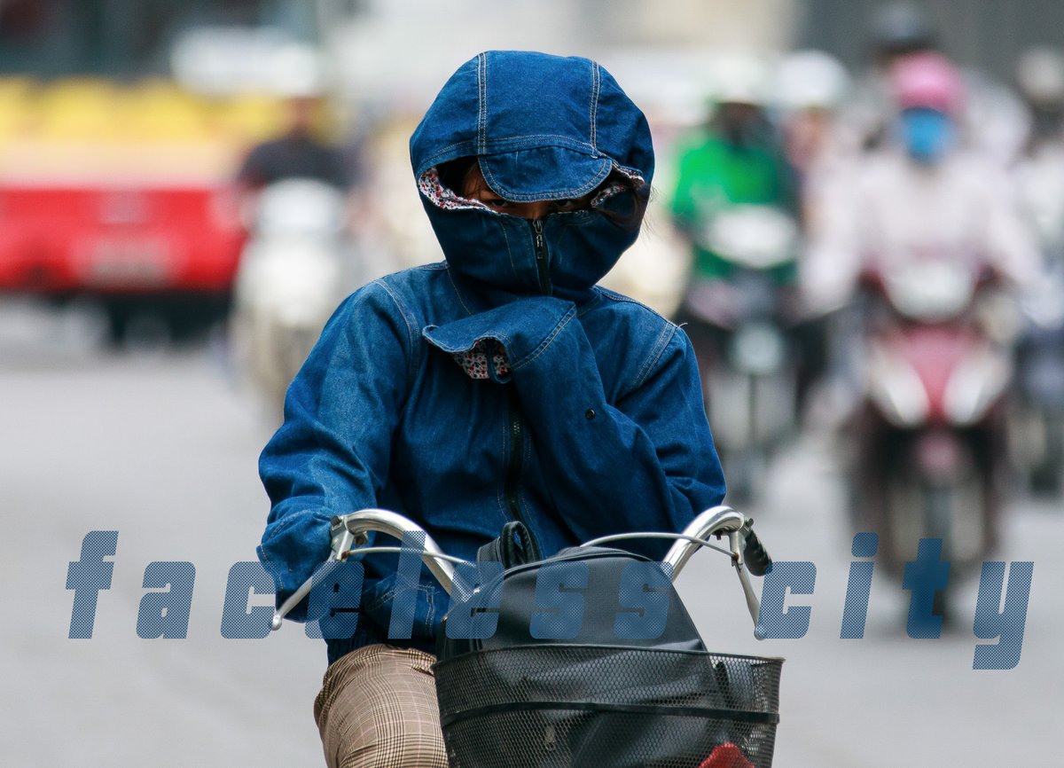 Bức ảnh chụp một người đi xe máy mặc trang phục bảo hộ kín mít, che mặt bằng khẩu trang và mũ trùm trong bối cảnh đường phố đông đúc. Hình ảnh tạo cảm giác về sự bảo vệ cá nhân trong môi trường ô nhiễm và giao thông tấp nập.