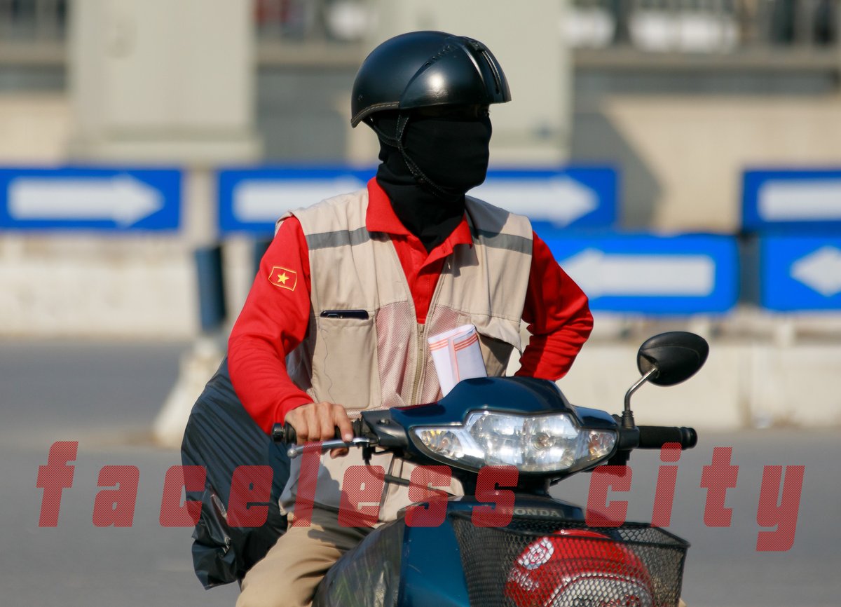 Hình ảnh một người đi xe máy trong thành phố, mặc áo đỏ và đội mũ bảo hiểm cùng khẩu trang che kín mặt. Cảnh vật thể hiện sự yên tĩnh và tập trung trong một khoảnh khắc giao thông đô thị.