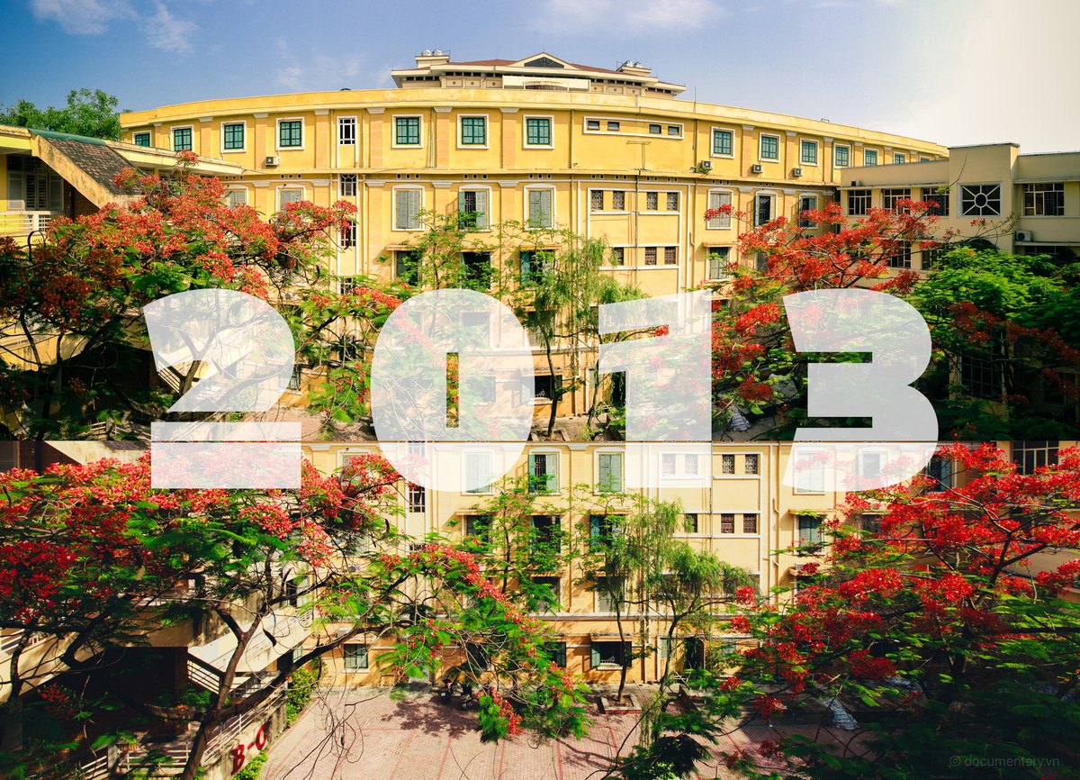 Bức ảnh chụp khuôn viên trường đại học với toà nhà màu vàng và cây phượng đỏ rực rỡ. Không gian tạo cảm giác yên bình và tươi sáng dưới ánh nắng ban ngày.