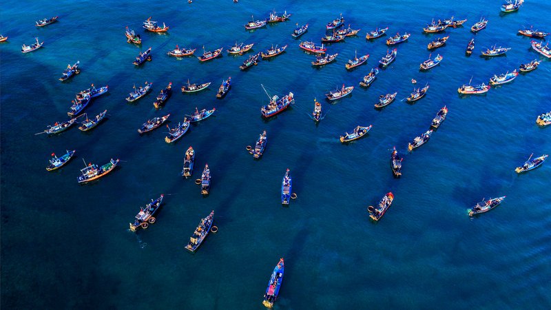 Hình ảnh toàn cảnh trên không của một làng chài với nhiều thuyền đánh cá trên biển xanh. Cảnh biển rộng lớn và yên bình tạo cảm giác thư thái và tĩnh lặng.