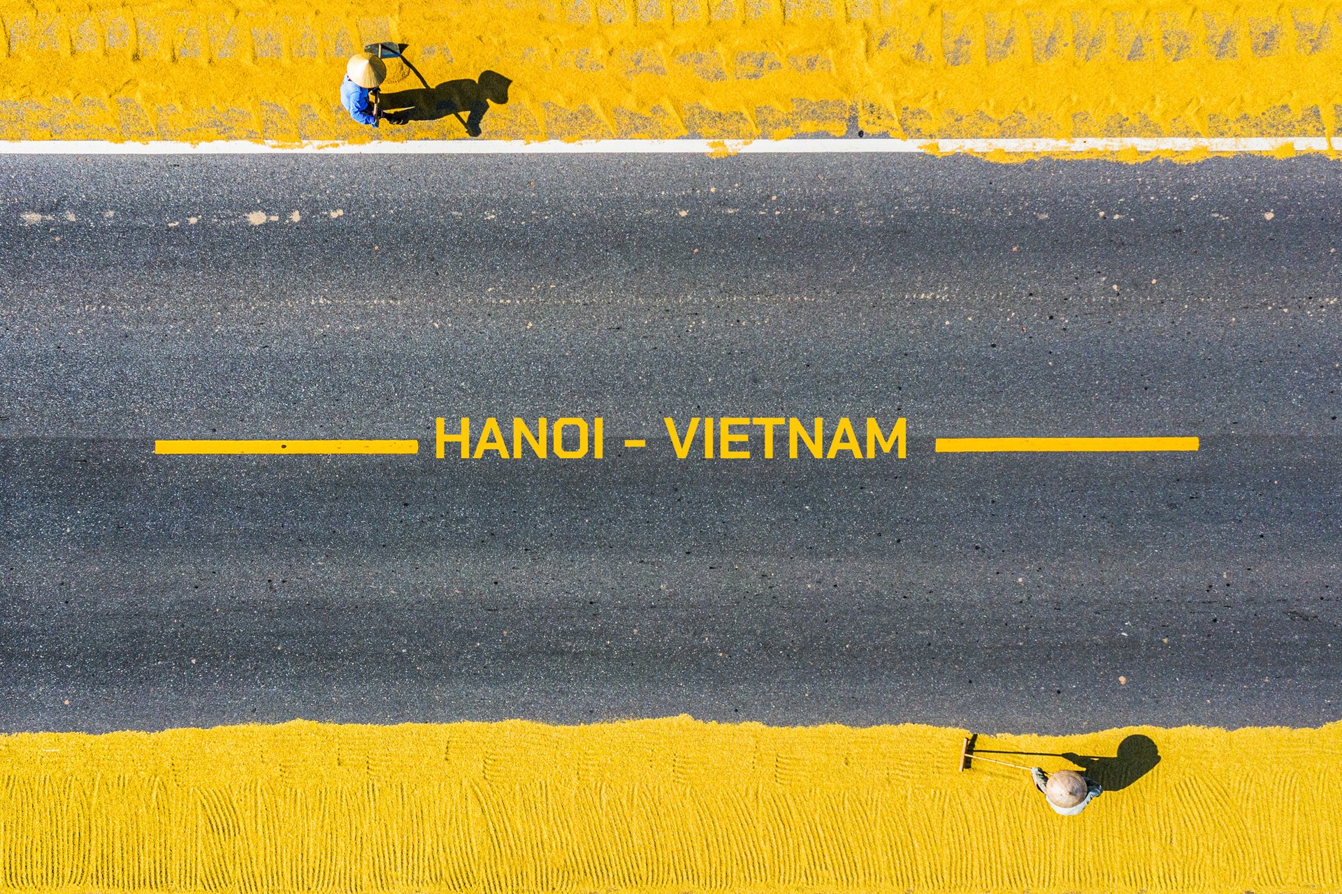 Bức ảnh chụp từ trên cao cho thấy cảnh phơi thóc trên hai bên đường nhựa với hai người đang làm việc. Hình ảnh tạo cảm giác yên bình và thể hiện nét đẹp trong cuộc sống nông thôn kết hợp với đô thị.