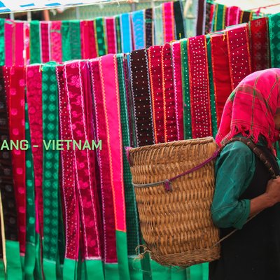 Bức ảnh chụp một người phụ nữ trong trang phục dân tộc tại chợ Hà Giang với các dải vải nhiều màu sắc treo phía sau. Hình ảnh thể hiện sự chân thực và sống động của đời sống thường nhật nơi đây.