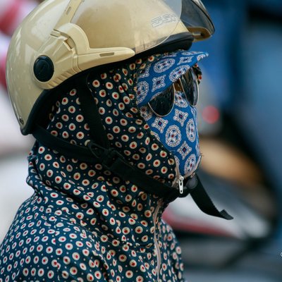 Hình ảnh một người mặc áo chống nắng hoạ tiết kín mít, đội mũ bảo hiểm và đeo khẩu trang cùng kính râm trên đường phố. Bức ảnh tạo cảm giác về sự bảo vệ cá nhân trong môi trường ô nhiễm và đông đúc.