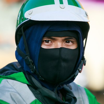 Bức ảnh chụp cận cảnh một người đi xe máy đội mũ bảo hiểm và đeo khẩu trang trong môi trường đô thị. Hình ảnh tạo cảm giác yên tĩnh và tập trung vào cá nhân trong không gian giao thông đông đúc.