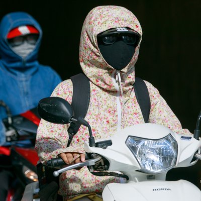 Hình ảnh hai người đi xe máy mặc áo chống nắng và khẩu trang kín mít trên đường phố. Cảnh tượng tạo cảm giác kín đáo và tập trung vào việc bảo vệ khỏi ánh nắng và bụi bẩn.