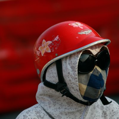 Bức ảnh chụp một người đội mũ bảo hiểm đỏ, đeo kính râm và khẩu trang, mặc áo chống nắng hoa văn trên phố. Hình ảnh tạo cảm giác bình yên và tập trung vào chi tiết trang phục bảo hộ cá nhân trong cuộc sống đô thị.