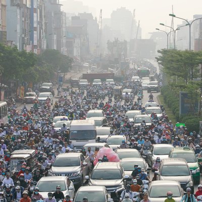 Hình ảnh cho thấy cảnh kẹt xe nghiêm trọng vào buổi sáng tại một ngã tư đông đúc với nhiều xe hơi và xe máy. Không khí có vẻ bị ô nhiễm và bụi mù bao phủ, tạo cảm giác ngột ngạt và bức bối trong thành phố.