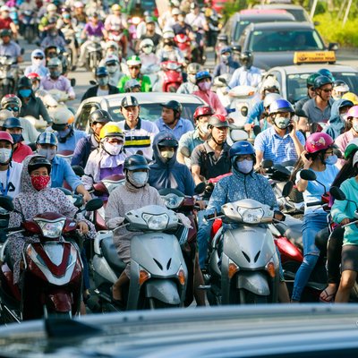 Bức ảnh chụp cảnh đông người đi xe máy và ô tô đang chờ đèn đỏ trên đường phố thành phố vào ban ngày. Hình ảnh thể hiện sự nhộn nhịp và tấp nập của giao thông đô thị với nhiều người đội mũ bảo hiểm và đeo khẩu trang.