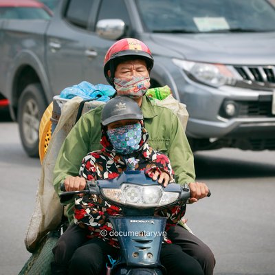 Hình ảnh hai người đi xe máy trên đường phố thành phố với ánh sáng tự nhiên và các phương tiện giao thông xung quanh. Bức ảnh thể hiện cảnh sinh hoạt thường nhật với cảm giác chân thực và gần gũi.