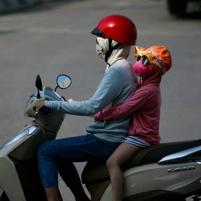 Bức ảnh chụp hai người trên một chiếc xe máy trong môi trường đô thị, cả hai đều mặc đồ bảo hộ và đeo khẩu trang để chống ô nhiễm. Cảm giác chung là sự bình yên trong cuộc sống thường nhật giữa nhịp sống đô thị sôi động.