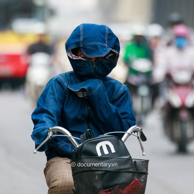 Hình ảnh một người đi xe máy mặc áo chống nắng và đeo khẩu trang kín mặt trên đường phố thành phố. Bức ảnh tạo cảm giác về sự bảo vệ cá nhân trong môi trường đô thị ô nhiễm và đông đúc.