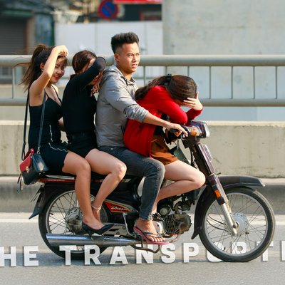 Bức ảnh chụp một nhóm bốn người đang đi trên một chiếc xe máy Honda Dream trên đường phố thành phố. Hình ảnh thể hiện sự đông đúc và vui nhộn trong giao thông đô thị vào buổi chiều.