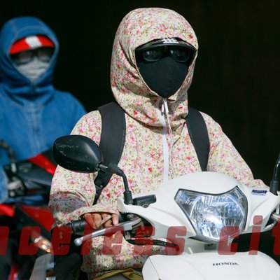 Hình ảnh hai người mặc áo chống nắng, đeo khẩu trang kín mít đang lái xe máy trên đường phố. Bức ảnh tạo cảm giác yên tĩnh và kín đáo trong một khung cảnh đô thị nhộn nhịp.