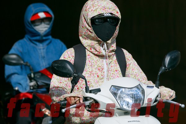 Hình ảnh hai người mặc áo chống nắng, đeo khẩu trang kín mít đang lái xe máy trên đường phố. Bức ảnh tạo cảm giác yên tĩnh và kín đáo trong một khung cảnh đô thị nhộn nhịp.