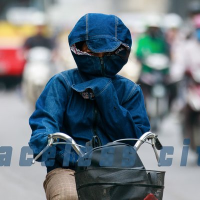 Bức ảnh chụp một người đi xe máy mặc trang phục bảo hộ kín mít, che mặt bằng khẩu trang và mũ trùm trong bối cảnh đường phố đông đúc. Hình ảnh tạo cảm giác về sự bảo vệ cá nhân trong môi trường ô nhiễm và giao thông tấp nập.