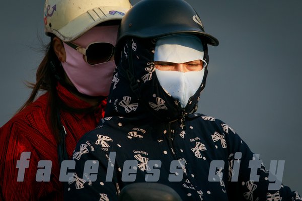 Bức ảnh chụp hai người trên xe máy trong trang phục bảo hộ với khẩu trang và mũ bảo hiểm, thể hiện cảnh đi lại trong thành phố. Cảm giác tổng thể là sự an toàn và bảo vệ trong môi trường đô thị có ô nhiễm.