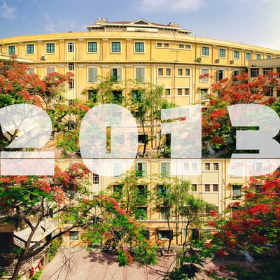 Bức ảnh chụp khuôn viên trường đại học với toà nhà màu vàng và cây phượng đỏ rực rỡ. Không gian tạo cảm giác yên bình và tươi sáng dưới ánh nắng ban ngày.