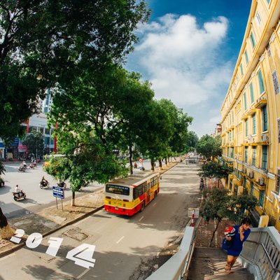 Hình ảnh một con đường ở Hà Nội với các cây xà cừ xanh mát, xe buýt và người đi bộ trên cầu thang. Không khí buổi sáng yên bình với ánh sáng nhẹ nhàng tạo cảm giác thư thái và sống động.