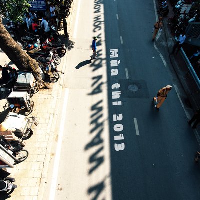 Bức ảnh chụp một con đường đông người với nhiều xe máy và cảnh sát giao thông đang làm nhiệm vụ. Không khí sôi động và căng thẳng của mùa thi được thể hiện rõ qua cảnh quan và hoạt động trên đường phố.