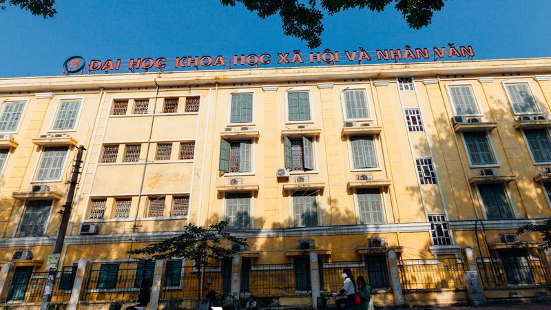 Bức ảnh chụp một toà nhà đại học tại Hà Nội với người đi xe đạp trên đường phố phía trước. Cảnh vật tạo cảm giác yên bình và sinh động của cuộc sống đô thị buổi sáng.