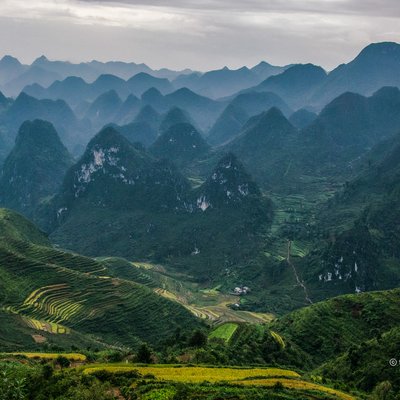 Bức ảnh chụp cảnh cao nguyên đá với các dãy núi và ruộng bậc thang xanh mướt dưới ánh sáng tự nhiên dịu nhẹ. Toàn cảnh mang lại cảm giác yên bình và hùng vĩ của thiên nhiên vùng cao nguyên.