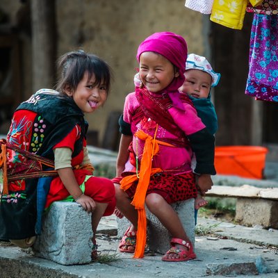 Bức ảnh chụp ba đứa trẻ mặc trang phục dân tộc truyền thống đang ngồi ngoài trời với ánh sáng tự nhiên. Cảnh tượng mang lại cảm giác vui tươi và sống động, thể hiện nét đẹp văn hoá và đời sống nông thôn vùng cao nguyên đá.