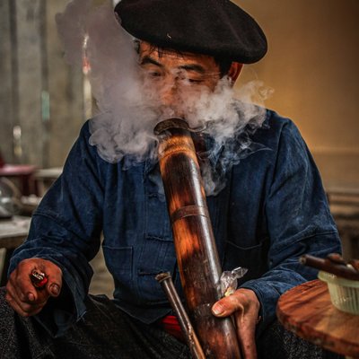 Bức ảnh chụp một người đàn ông đang hút thuốc lào bằng ống tre trong không gian trong nhà. Hình ảnh thể hiện phong tục truyền thống và đời sống thực của người dân vùng cao nguyên đá Việt Nam.
