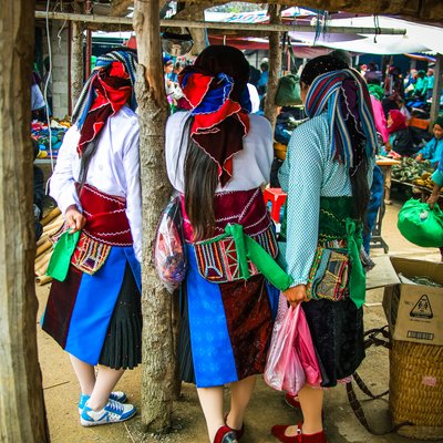 Bức ảnh chụp cảnh ba người phụ nữ mặc trang phục dân tộc truyền thống tại một chợ phiên ở vùng cao nguyên đá. Không khí sôi động và màu sắc sặc sỡ của trang phục tạo nên cảm giác sống động và đậm đà bản sắc văn hoá.